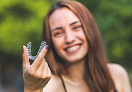 Woman with brown hair and purple nails outside holding clear aligner smiling